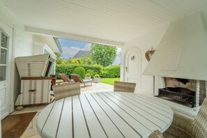 A patio with a white table and chairs, a fireplace, and a view of the garden. - Söns-rose - Luxusoase mit Traumhaften Garten in Munkmarsch am Wattenmeer (Sylt-Ost)