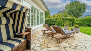 Eine Terrasse mit Steinplattenboden, einem gestreiften Sofa und Holzliegestühlen.