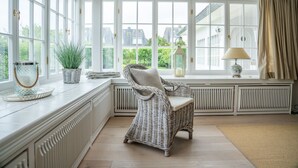 Ein helles Zimmer mit großen Fenstern, einem Korbstuhl, einem Heizkörper und einer Lampe.