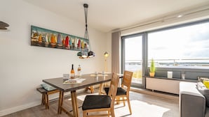 A modern dining area with a wooden table and chairs, a painting of sailboats on the wall, and a large window offering a view of the sea.