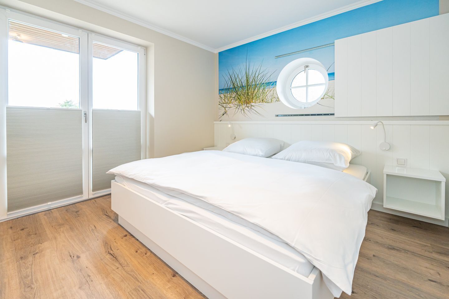 Ein Schlafzimmer mit einem großen Bett, einer Schiebetür zum Glas und Blick auf den Strand sowie einem runden Fenster.