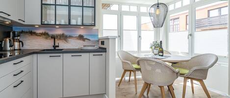 Une cuisine moderne avec des armoires blanches, un comptoir noir et un revêtement mural sur le thème de la plage. On y trouve un coin repas avec des chaises et une table en osier, ainsi que de grandes fenêtres équipées de stores.