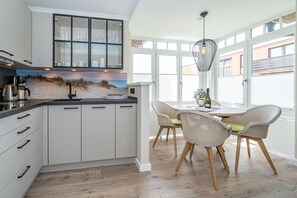 A modern kitchen with white cabinets, a black countertop, and a beach-themed backsplash. There is a dining area with wicker chairs and a table, and large windows with blinds.