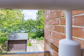 A view through a window of a brick wall and a wooden shed with a metal roof, overlooking a stone wall and lush green trees. - Strandperle - Modern 2-room vacation apartment with balcony, by the village pond. (Wenningstedt-Braderup (Sylt))