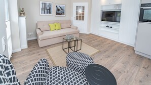 Una sala de estar moderna con un sofá, una mesa de centro y un otoman estampado. Se observa una puerta con ventana, una zona de cocina con televisión y horno, y una pared con cuadros enmarcados.