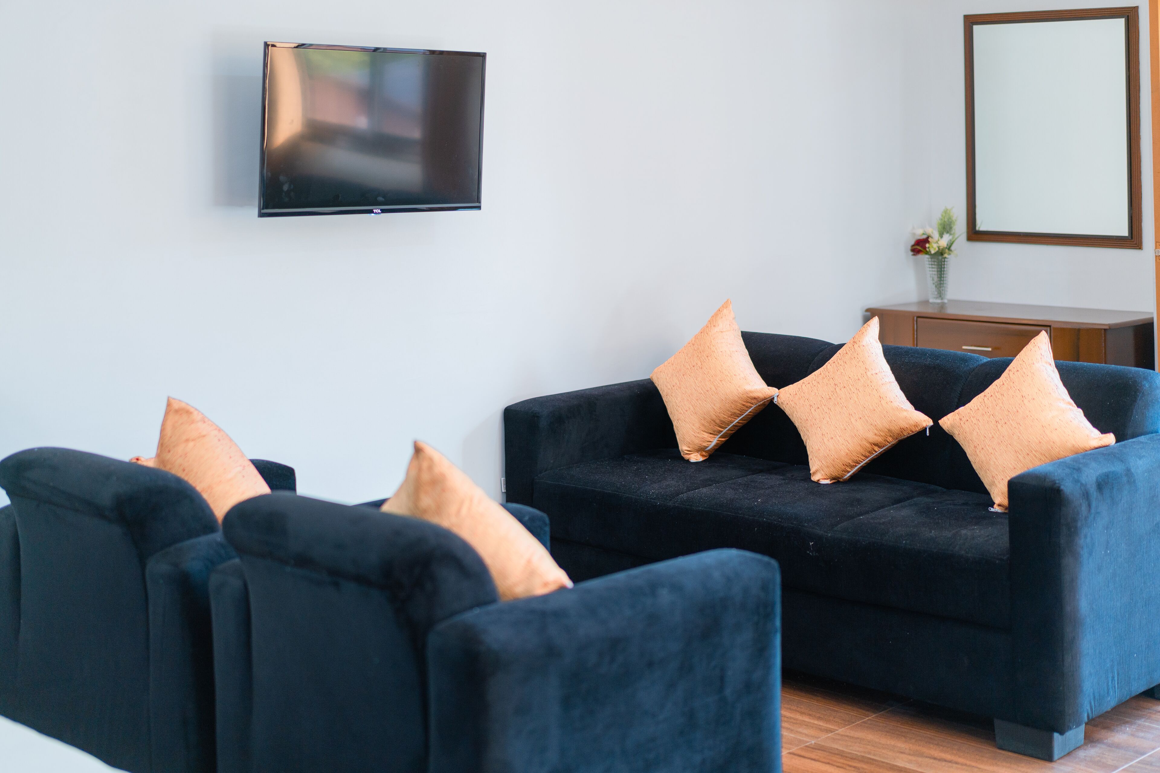 A modern living room with a dark blue sofa set, a flat-screen TV mounted on the wall, and a small wooden cabinet with a vase of flowers.