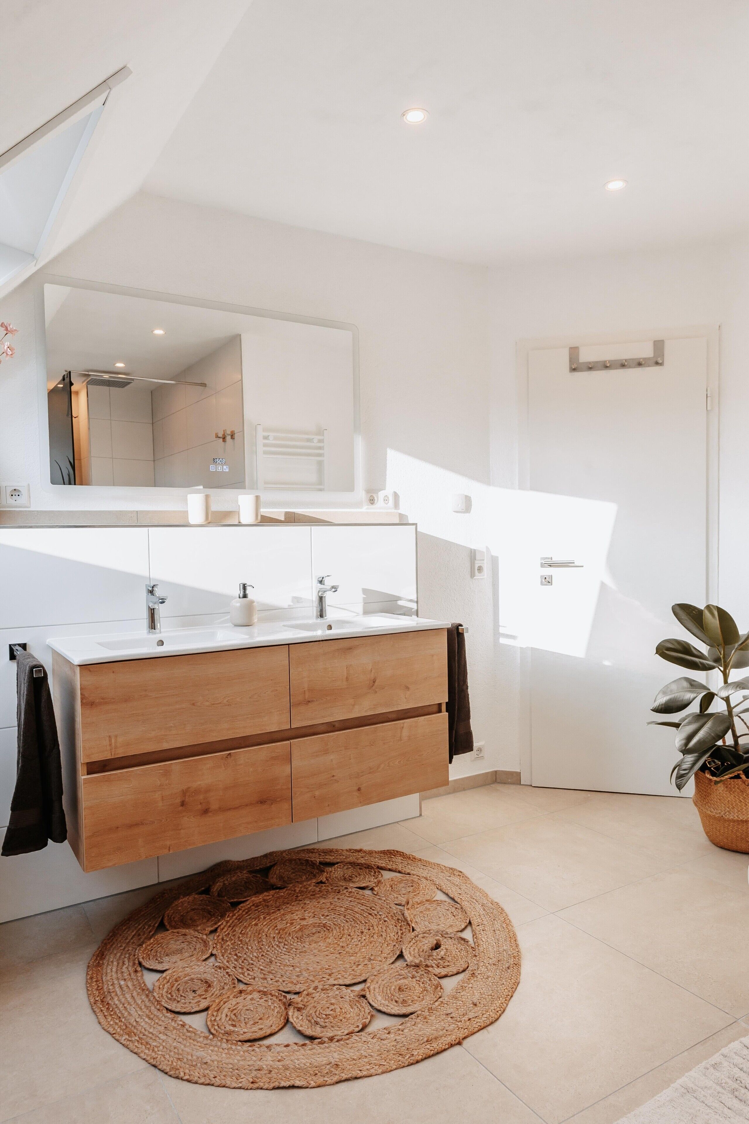 Een moderne badkamer met een houten wastafelmeubel, een rond geweven vloerkleed en een grote spiegel.