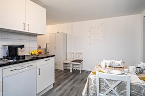 Una cocina moderna con armarios blancos, una cafetera y un frutero. Hay dos sillas blancas y una mesa de comedor con platos, vasos y un mantel estampado.