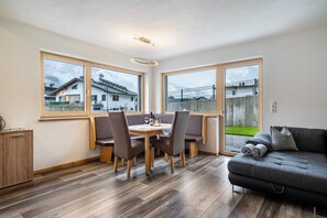 Ein moderner Essbereich mit einem Holztisch, Stühlen, großen Fenstern und einem grauen Sofa.