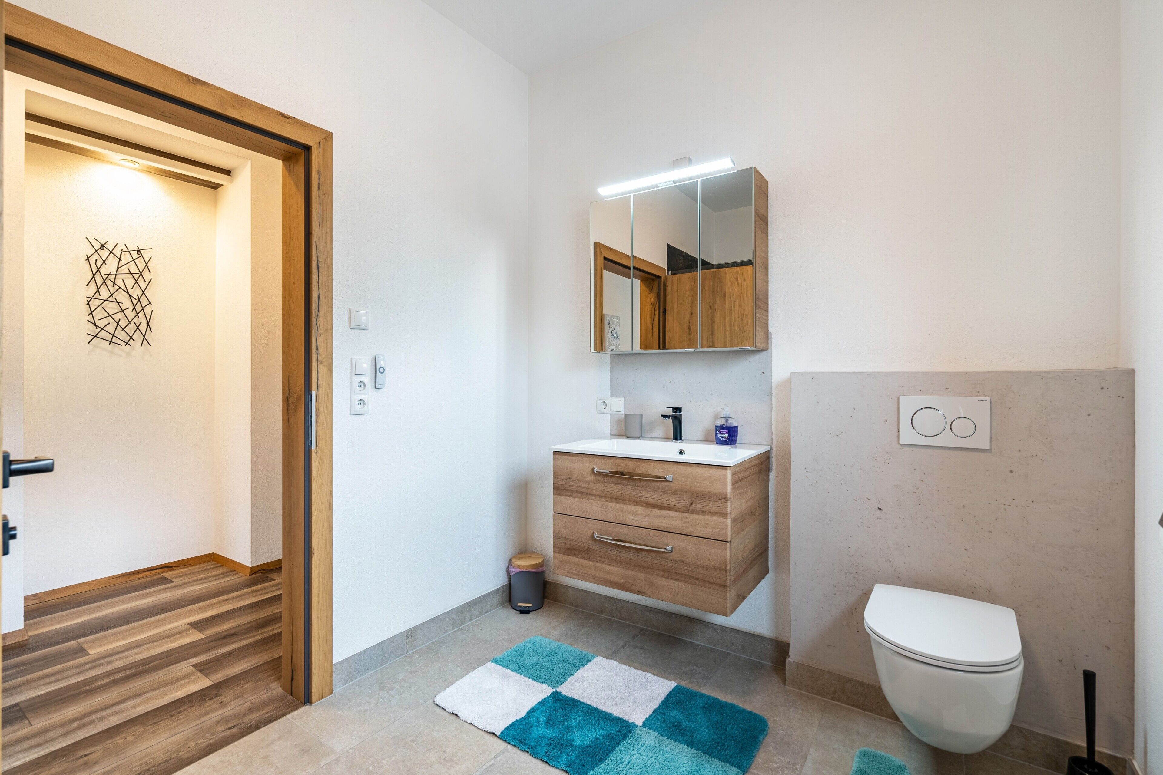 Een moderne badkamer met een houten wastafelmeubel, een spiegel, een wit toilet en een blauw-wit tapijt.