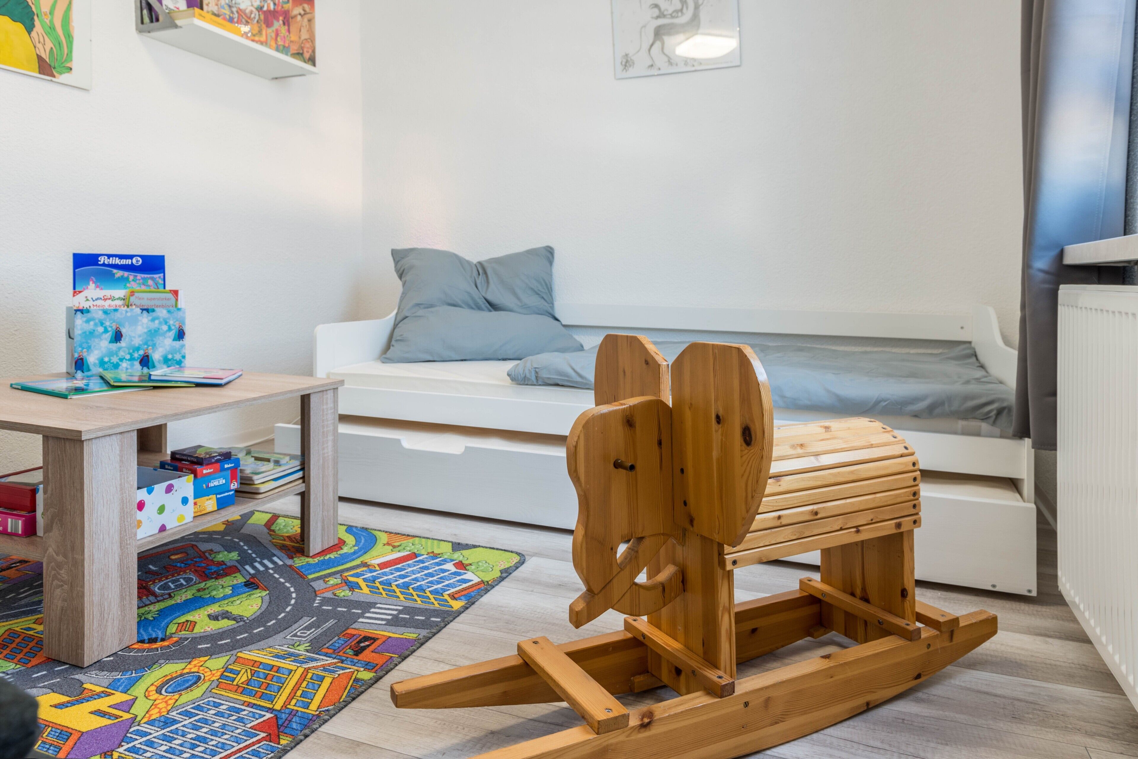 A child's playroom with a bed, a wooden rocking horse, a colorful play mat, and a shelf with books and toys.