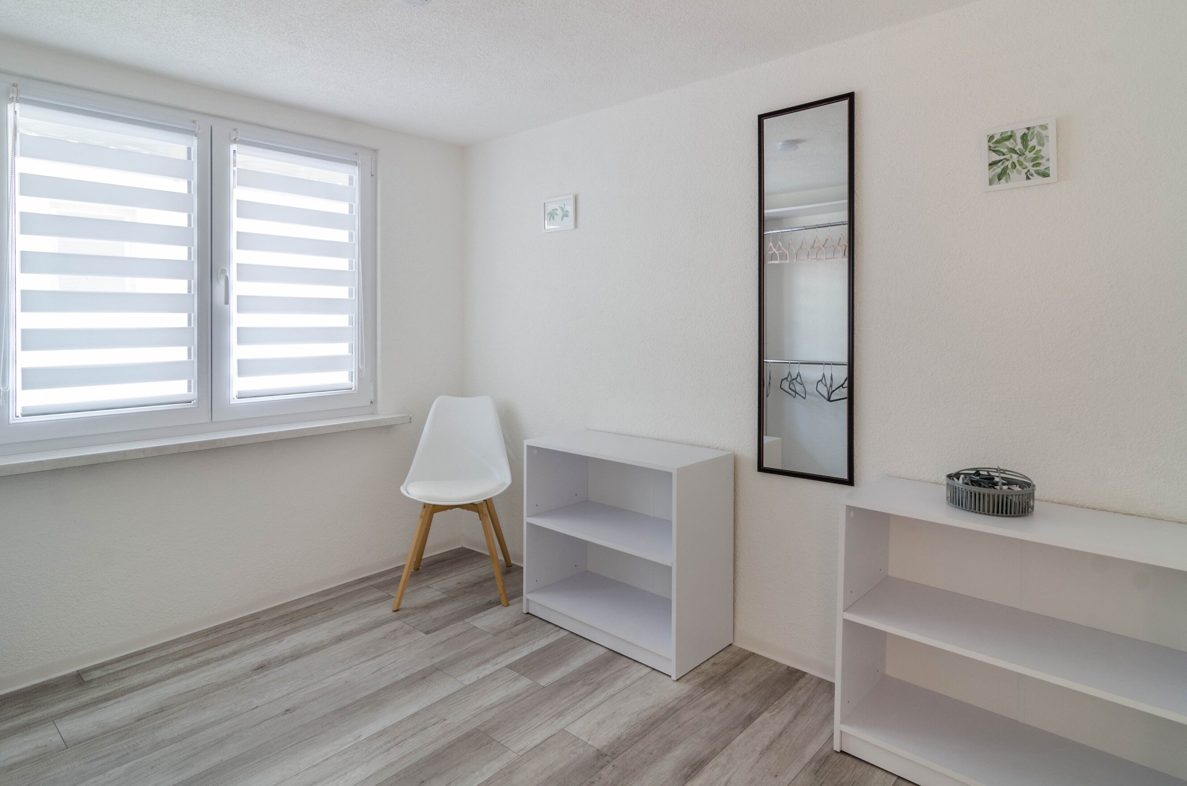 A room with a white chair, a white shelving unit, and a large mirror on the wall.