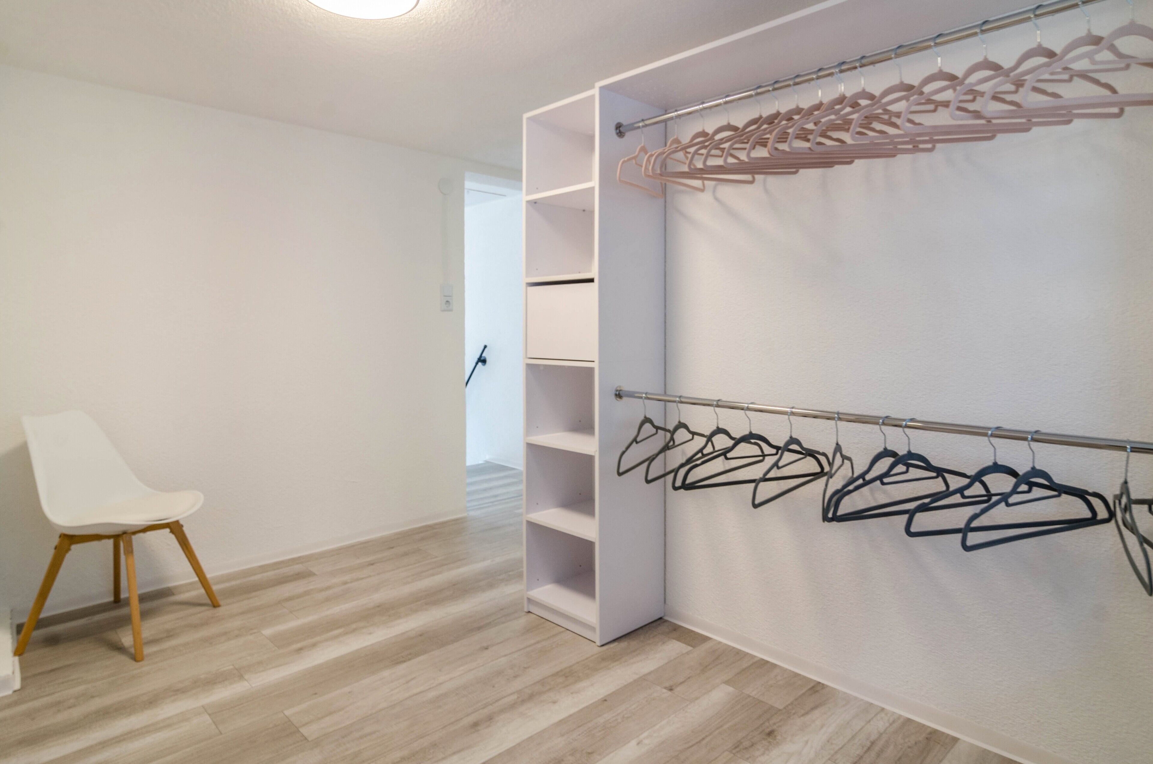 A room with a white chair, a closet with hanging clothes, and a wooden floor.