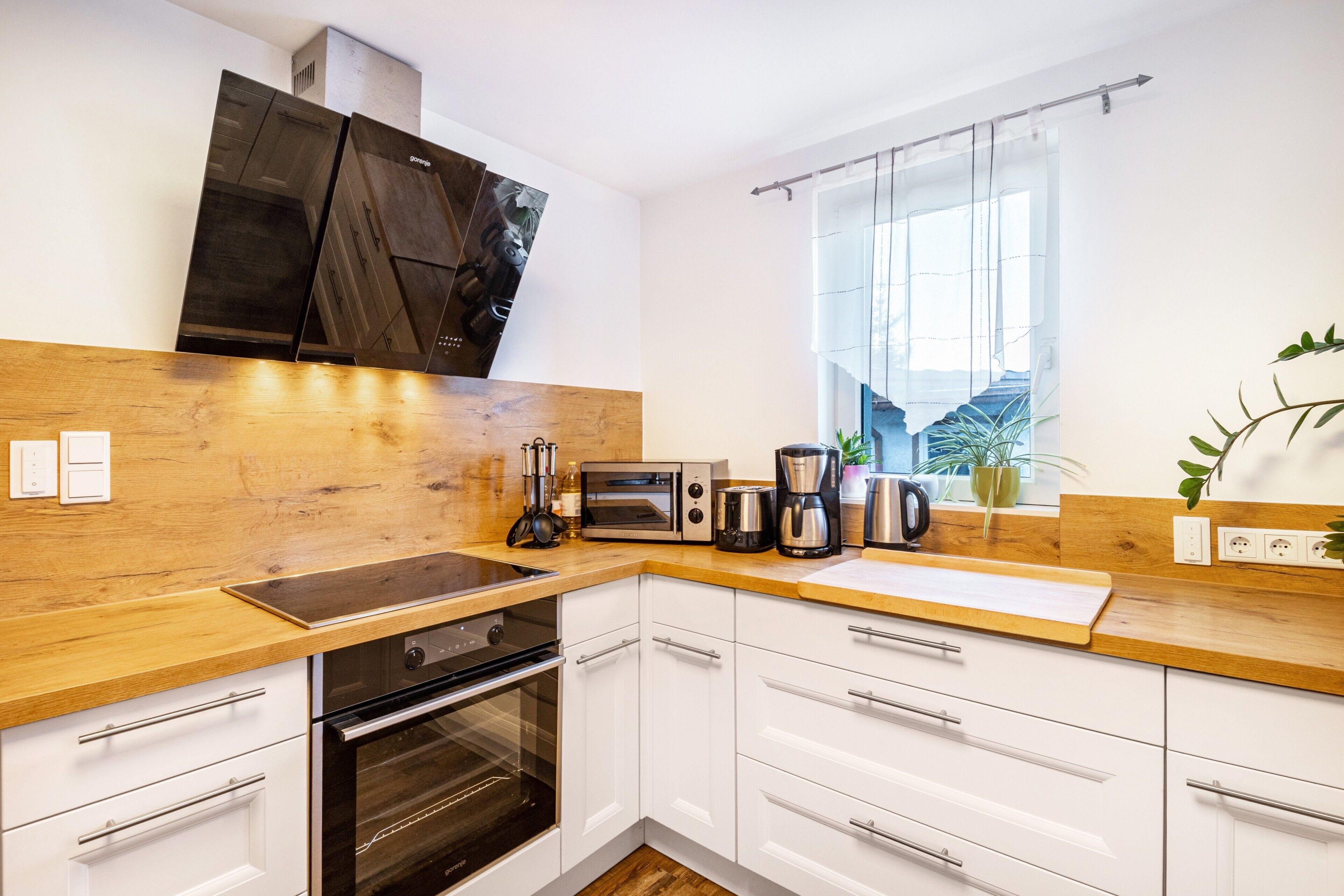 A modern kitchen with white cabinets, a wooden countertop, a built-in oven, a coffee maker, and a window with a view.