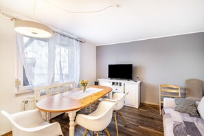A room with a wooden dining table, white chairs, a TV on a white cabinet, a window with sheer curtains, and a bed with a patterned blanket.