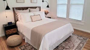 Een slaapkamer met een groot bed, twee nachtkastjes, een rond pouf, een getextureerd vloerkleed en twee wandlampen.