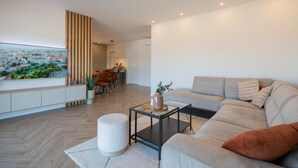 Ein modernes Wohnzimmer mit großem Flachbildfernseher, einer Couch, einem Couchtisch und einem Essbereich im Hintergrund.