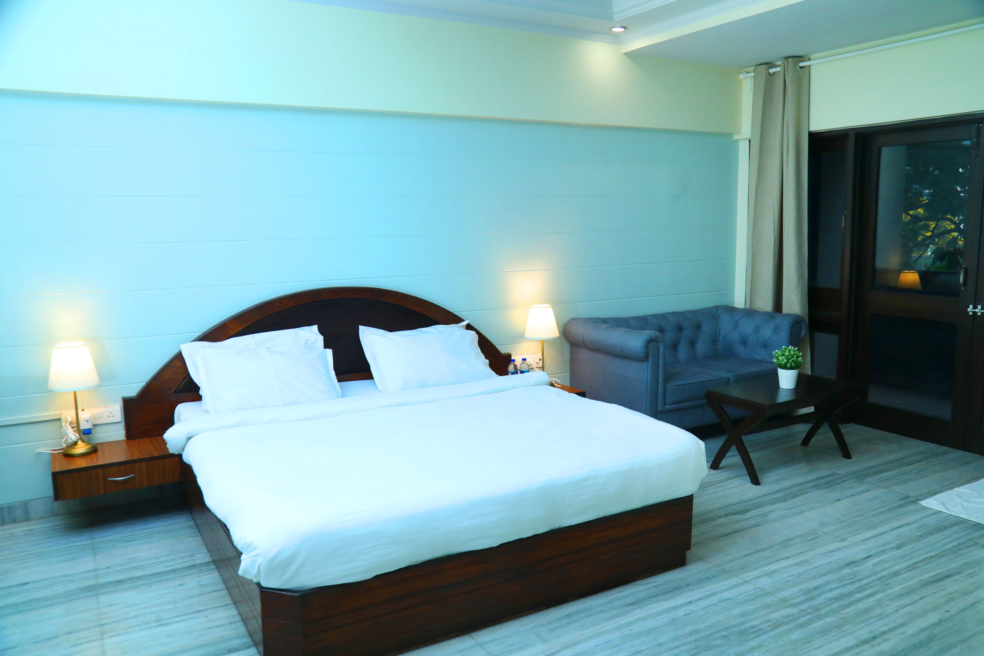 A hotel room with a large bed, two bedside lamps, a grey sofa, a small table with a plant, and a sliding glass door to the outside.