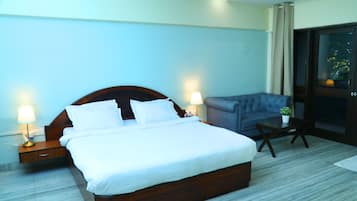 A hotel room with a large bed, two bedside lamps, a grey sofa, a small table with a plant, and a sliding glass door to the outside.