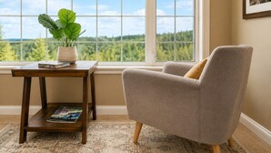 A room with a view through a large window, a grey armchair, a small wooden table with a plant and a magazine, and a framed picture on the wall. - Gigantic 6b5b home with stunning view & spacious rooms (Fairbanks)