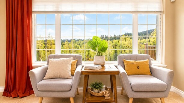 A room with two grey armchairs, a wooden table with a plant, and a view of a forest through large windows. - Gigantic 6b5b home with stunning view & spacious rooms (Fairbanks)
