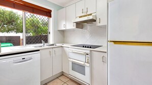 Een kleine keuken met witte kasten, een inbouwoven, een koelkast en een raam met zonwering.