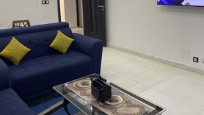 A modern living room with a blue sofa, a glass coffee table, and a flat-screen TV mounted on the wall. - SECKEBE IMMO ET BUSINESS: SUITE1 (DAKAR)