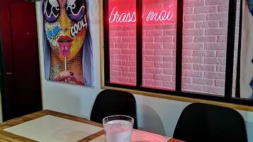A dining area with a wooden table, black chairs, a glass of milk, and a neon sign reading 'brass mai' on a brick wall.