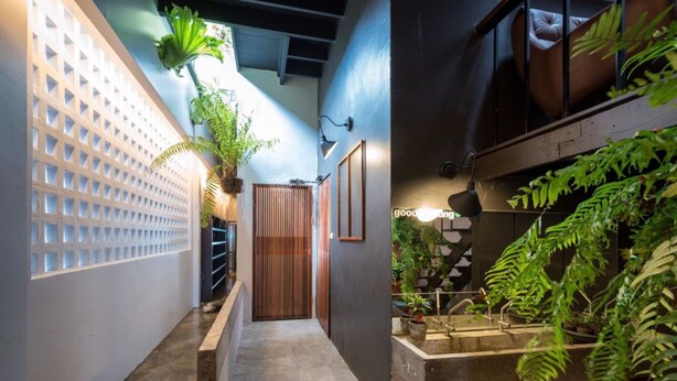 A narrow hallway with a concrete floor, a wooden door, and a wall with a geometric pattern. There are potted plants and a small fountain on the right side. - The Urban Room (Phuket)