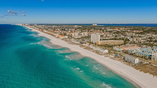 Luchtbeeld van een kustplaatsje met een lang zandstrand, helder turquoise water en een mix van woon- en bedrijfsgebouwen.