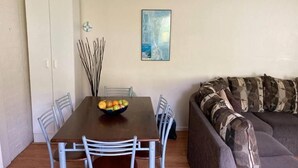 Un coin repas avec une table, des chaises et un panier de fruits. On y trouve également un meuble blanc, un canapé et un tableau accroché au mur.