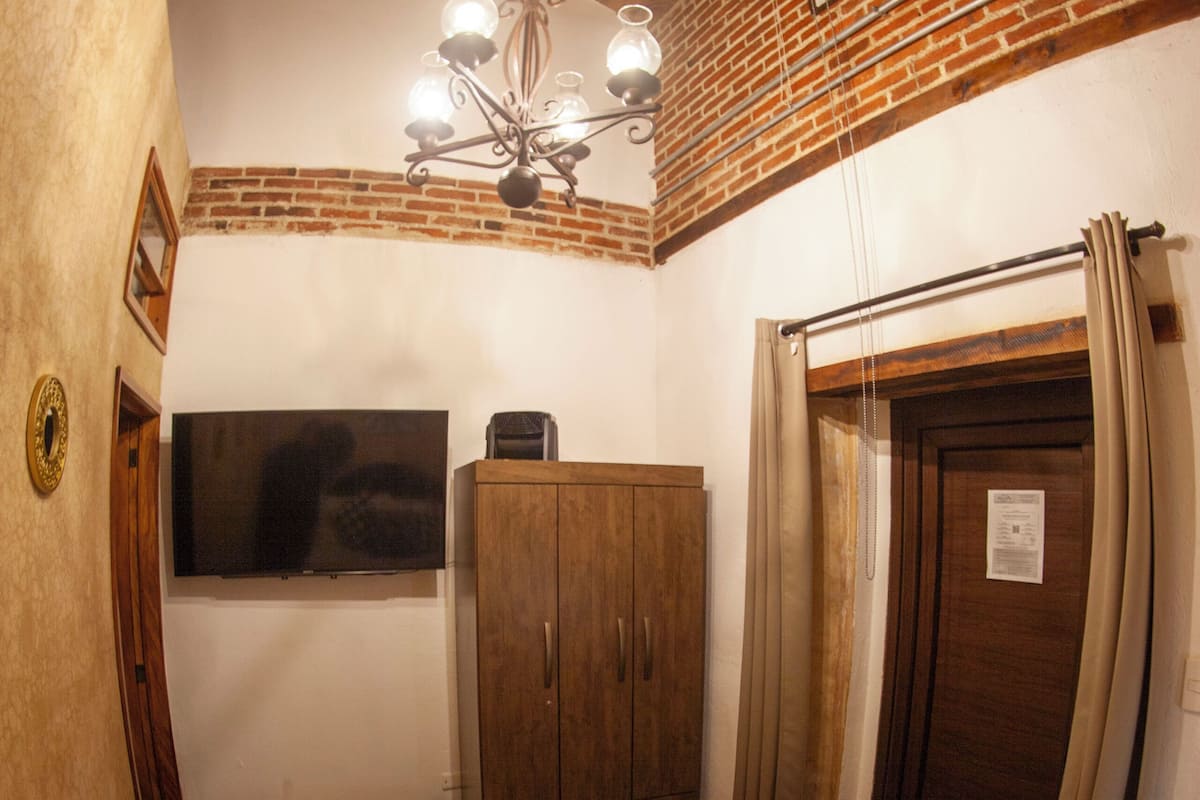 A bedroom with a bed, a wooden wardrobe, a television mounted on the wall, and a ceiling with exposed brick and wooden beams.