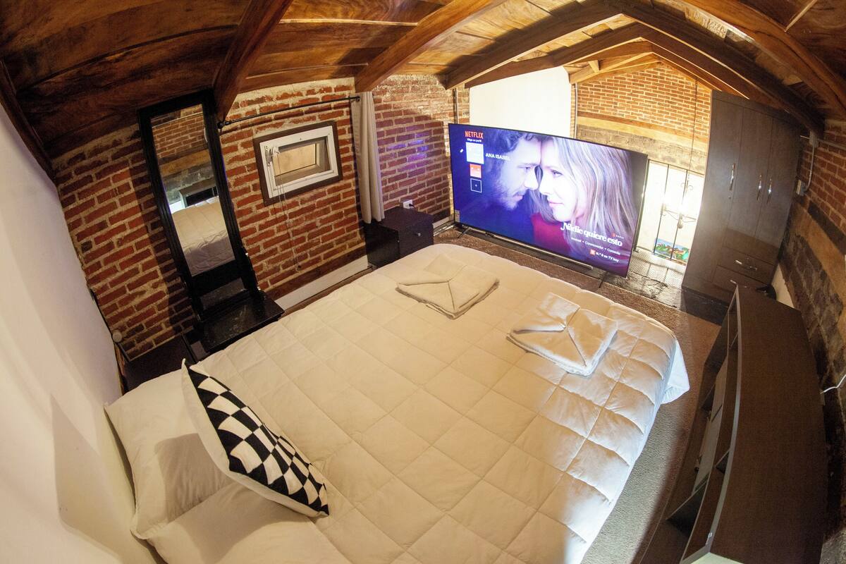 A bedroom with a brick wall, a large bed with a quilted cover, a flat-screen TV, and a wooden ceiling.