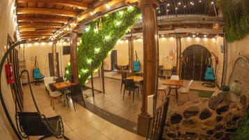 A cozy, rustic interior with wooden beams, stone walls, and a green moss-covered archway. There are multiple seating areas with tables and chairs, and a fire extinguisher is visible on the left.