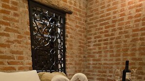 Habitación de hotel con pared de ladrillo, una reja metálica decorativa, una cama con pétalos de rosa, una botella de vino y copas sobre una mesita de noche.