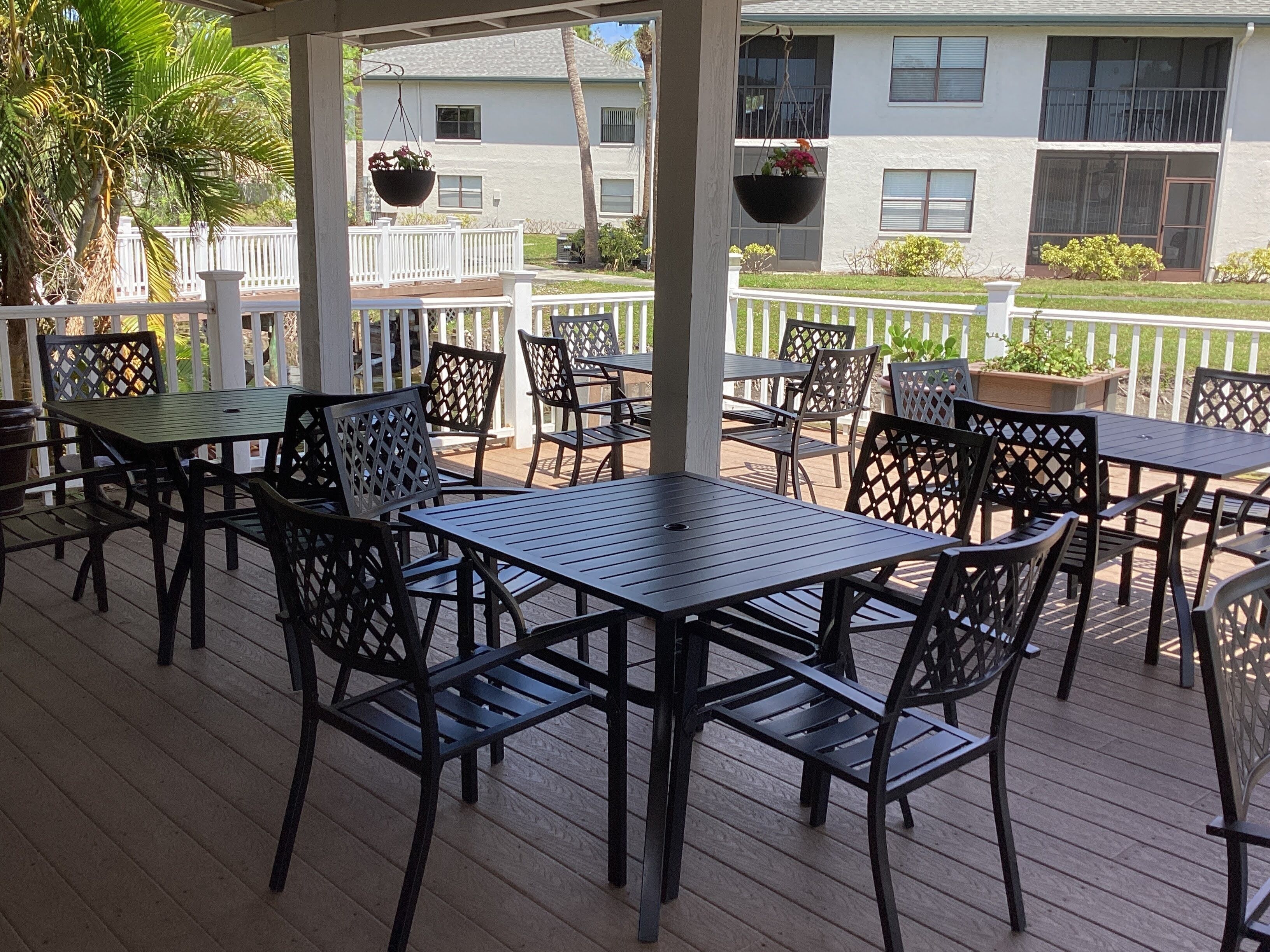 A covered outdoor patio with multiple tables and chairs, a white fence, and hanging flower baskets.