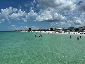 Ein Strand mit klarem türkisblauem Wasser, Menschen beim Schwimmen und Sonnenbaden, und Gebäuden im Hintergrund.