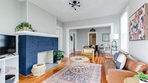 Ein Wohnzimmer mit blauem Kamin aus Ziegelsteinen, einem Fernseher, einem Couchtisch, einer Couch und einem Essbereich im Hintergrund.