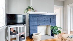 Ein Wohnzimmer mit Fernseher auf einem weißen Schrank, einem blauen Kamin aus Ziegeln und einem Couchtisch aus Weidengeflecht.