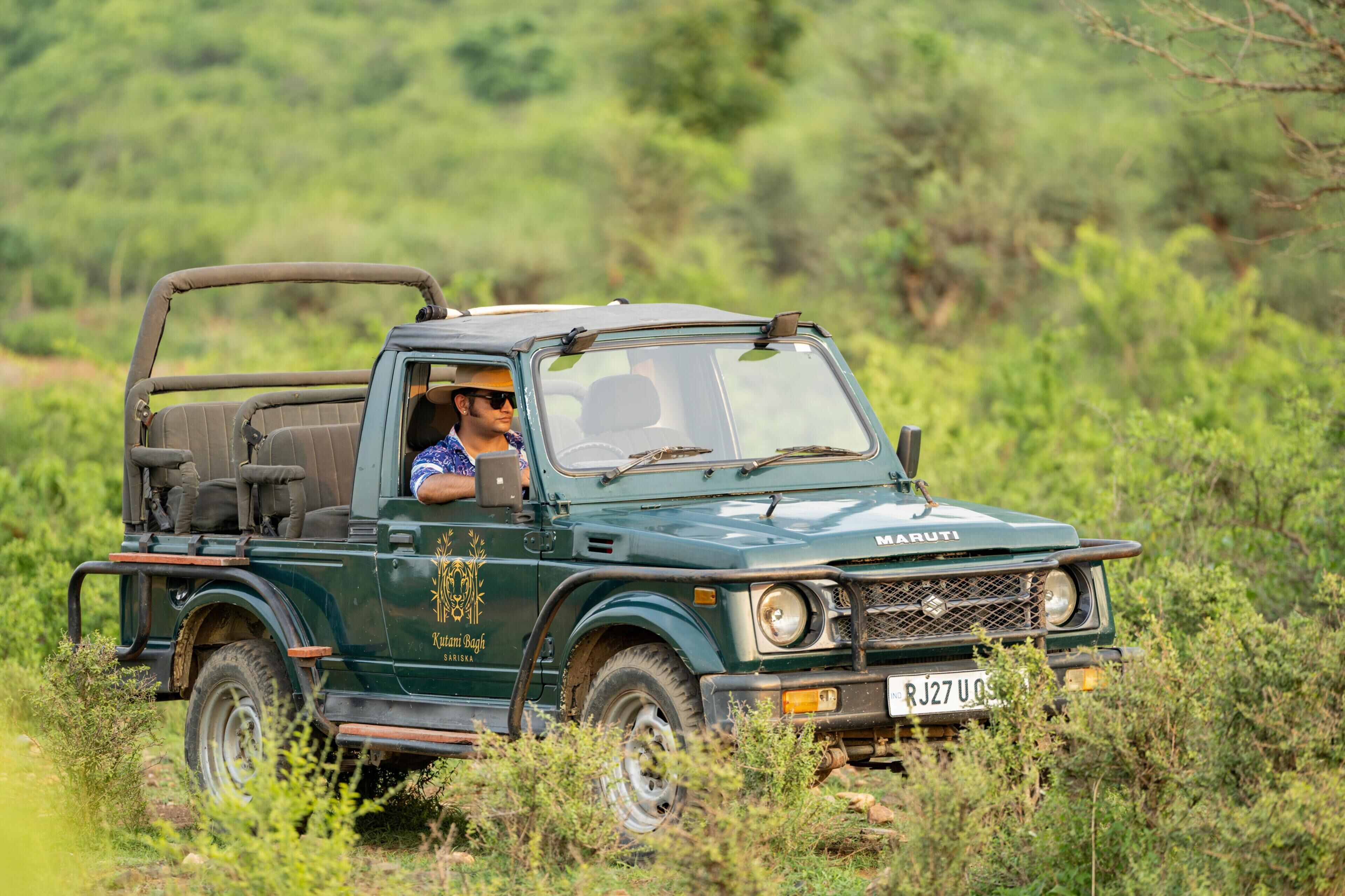 A green Maruti pickup truck with a spare tire mounted on the back, driving through a grassy area.