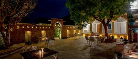Un patio exterior con suelo de piedra, arreglos de asientos y detalles arquitectónicos iluminados.