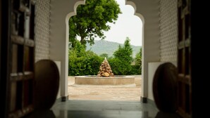 An arched doorway leading to a courtyard with a stone structure and a large tree.