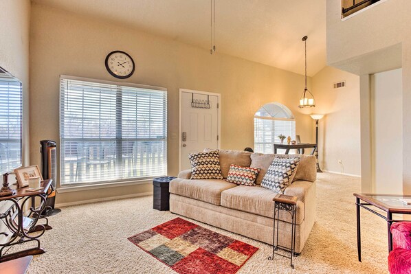 A living room with a beige sofa, patterned cushions, a red and gray rug, a clock on the wall, and a large window with blinds.