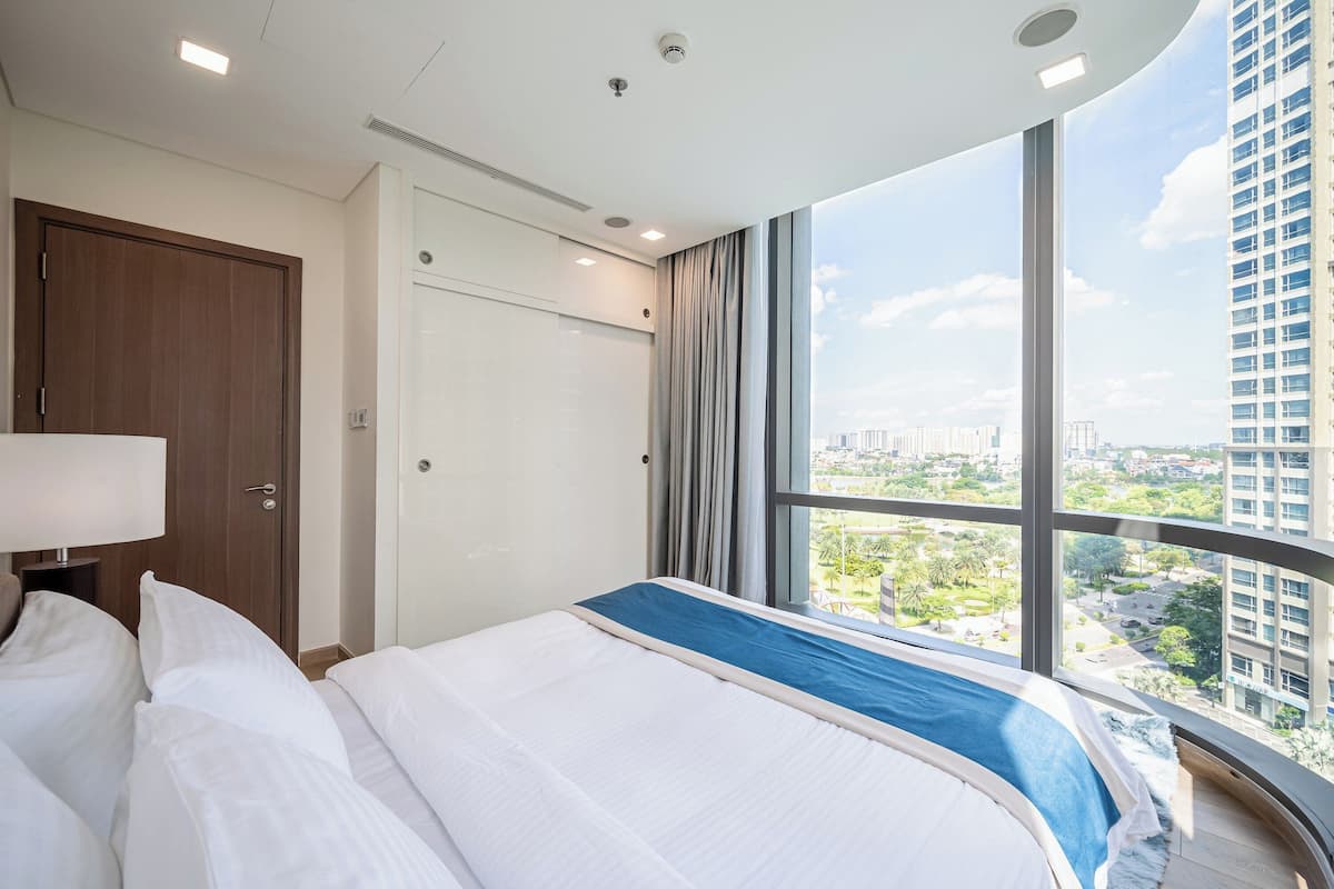 Habitación de hotel con una cama grande, una mesita de noche, una lámpara, vistas de la ciudad a través de amplias ventanas y una puerta.