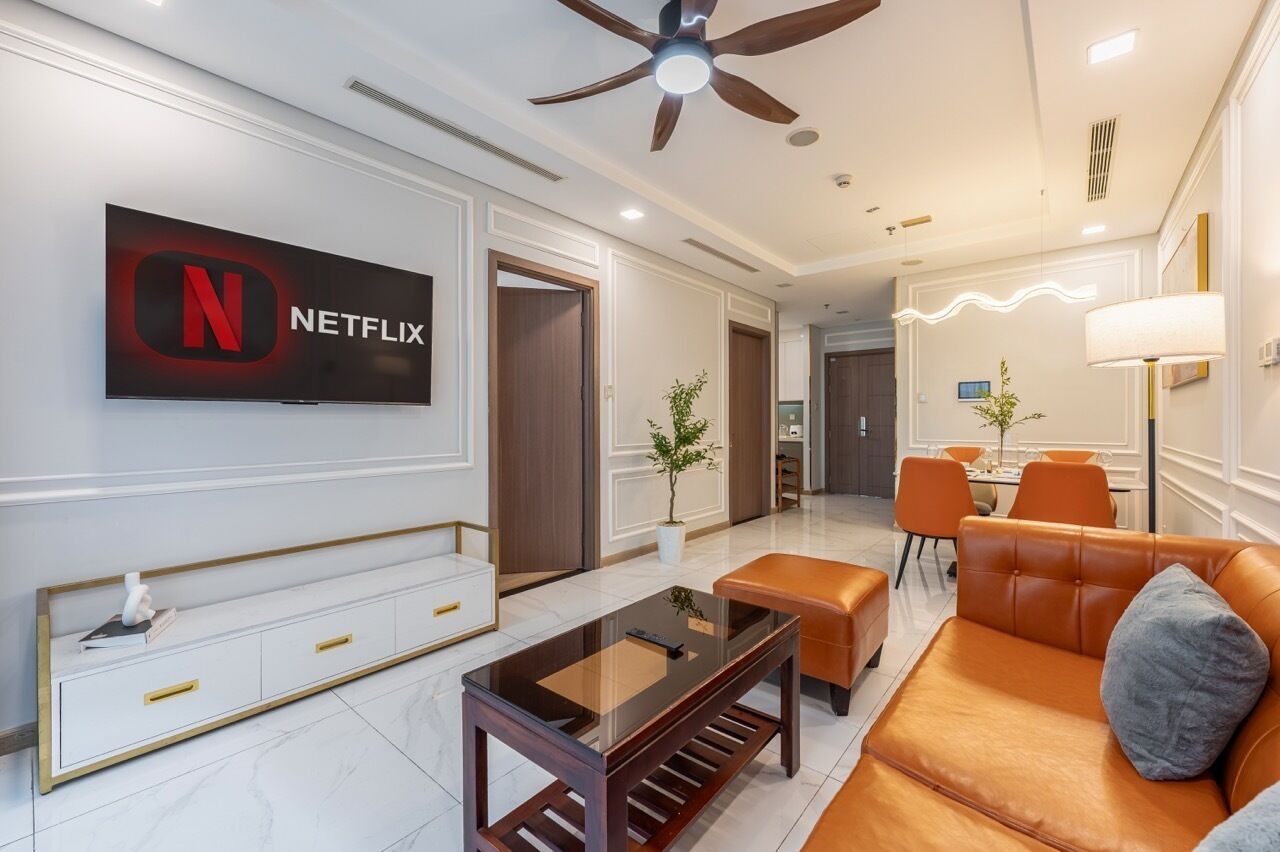 A modern living room with a Netflix logo, a ceiling fan, a glass coffee table, a leather sofa, and a dining area with orange chairs.