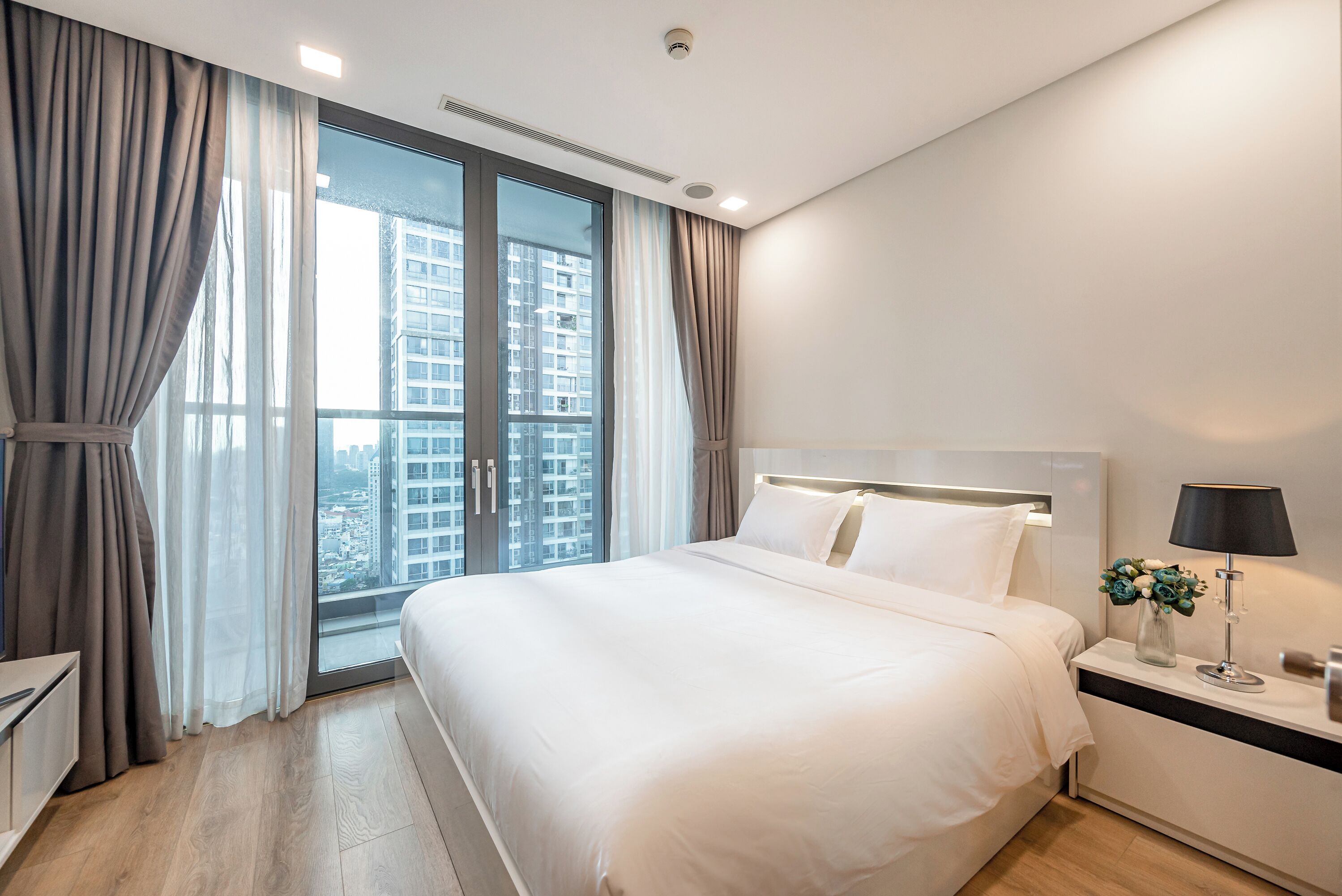 A modern hotel room with a large bed, a bedside table with a lamp, and a view of a cityscape through the window.