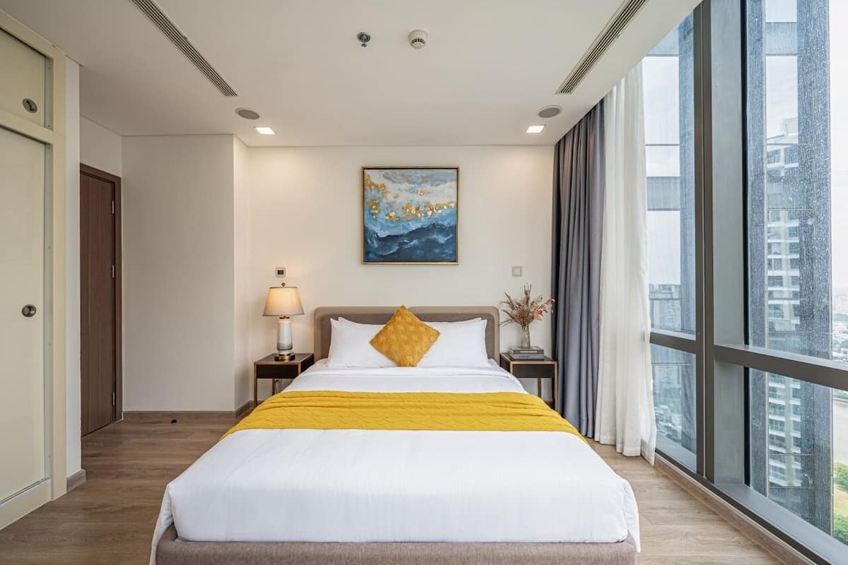 Habitación de hotel con una cama grande, una mesita de noche con lámpara, un cuadro en la pared y vistas de edificios a través de la ventana.