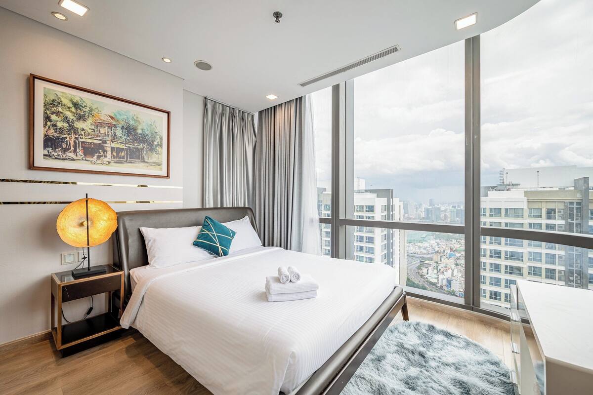 Una habitación de hotel moderna con una cama grande, una mesita de noche con lámpara, vistas de la ciudad a través de grandes ventanales y un cuadro en la pared.