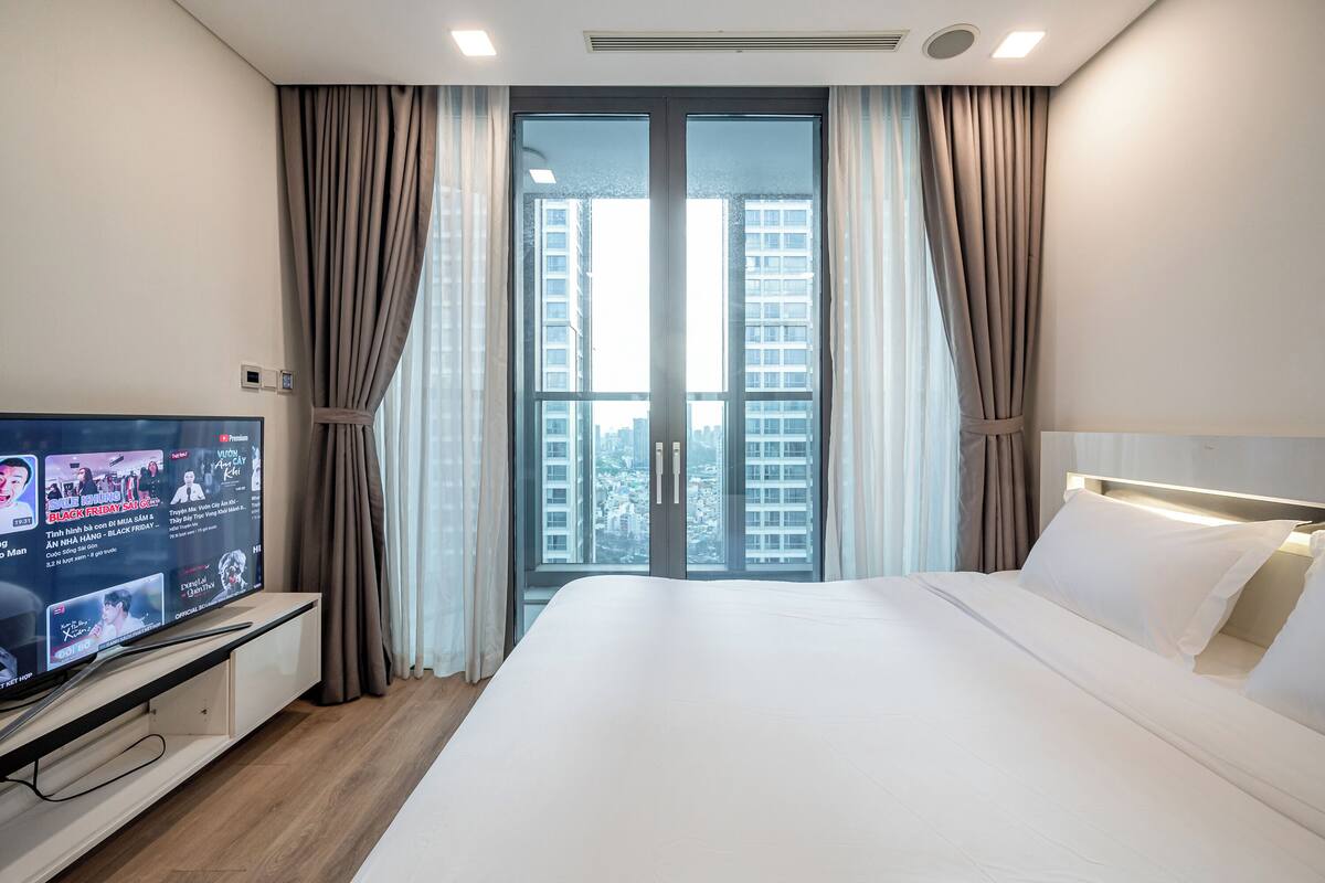 Habitación de hotel con una cama grande, un televisor sobre un pedestal y vistas a edificios altos por la ventana.