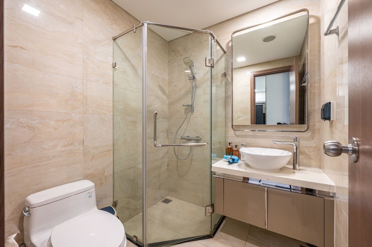Un baño moderno con ducha de vidrio, inodoro blanco, lavamanos con espejo y paredes de azulejos color beige.